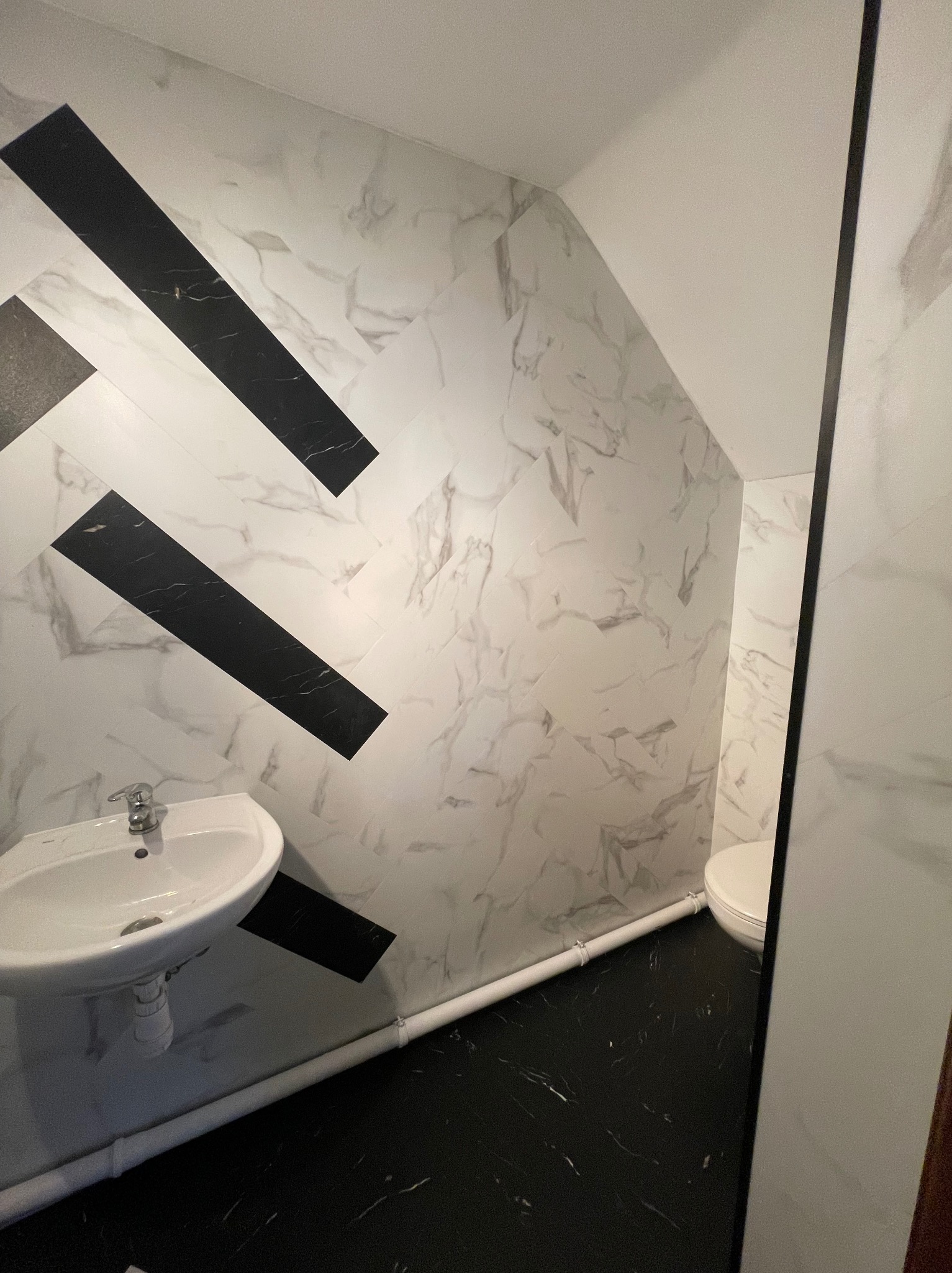 Wykończona łazienka z czarną podłogą imitującą marmur i ścianami wyłożonymi białymi płytkami z szarym marmurkowym wzorem, ułożonymi w geometryczny wzór z czarnymi akcentami, widoczna biała umywalka...