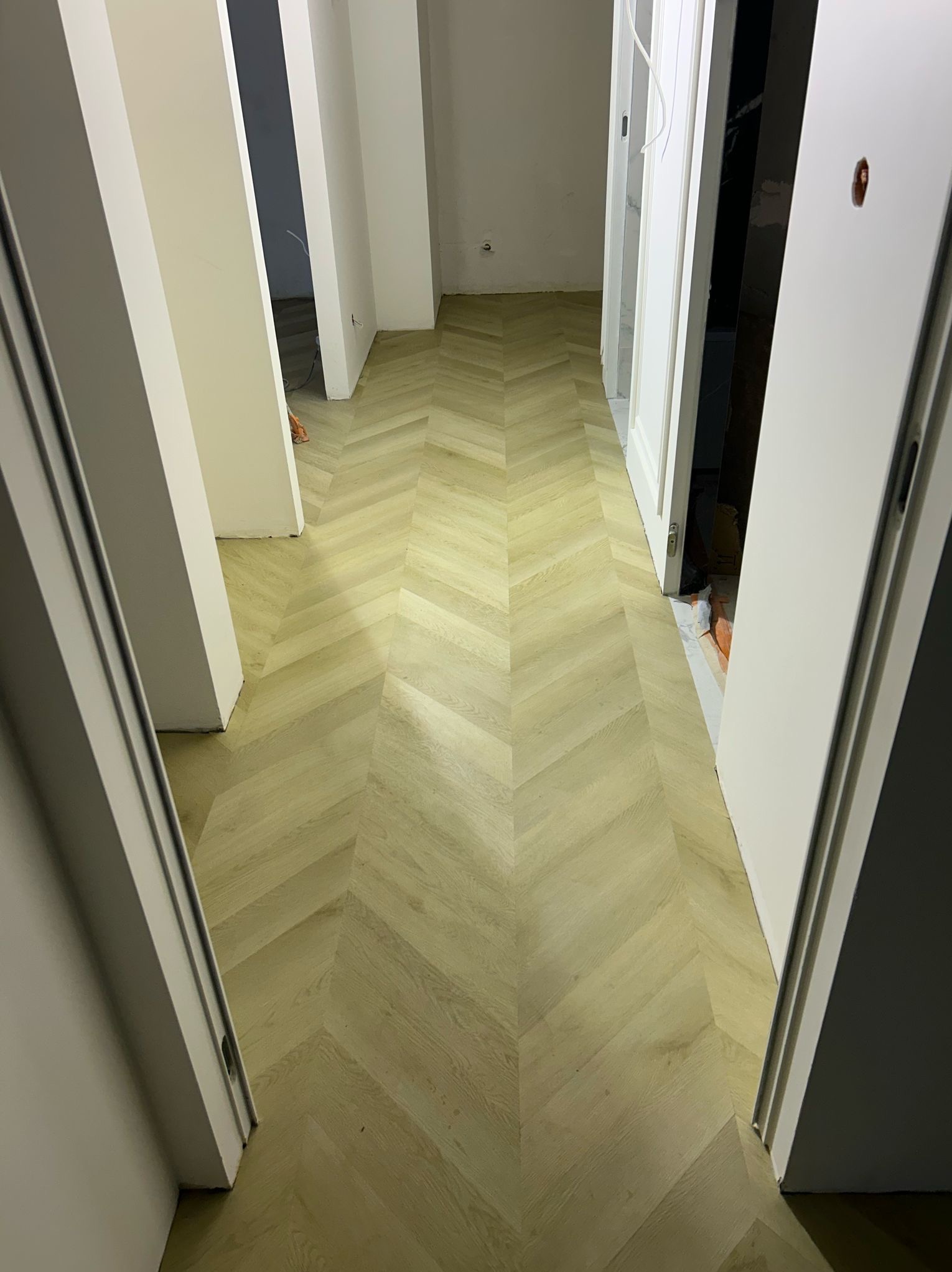 Jasna, nowa podłoga ułożona we wzór jodełki w korytarzu, widok zza otwartych drzwi, częściowo widoczne inne pomieszczenia w tle.