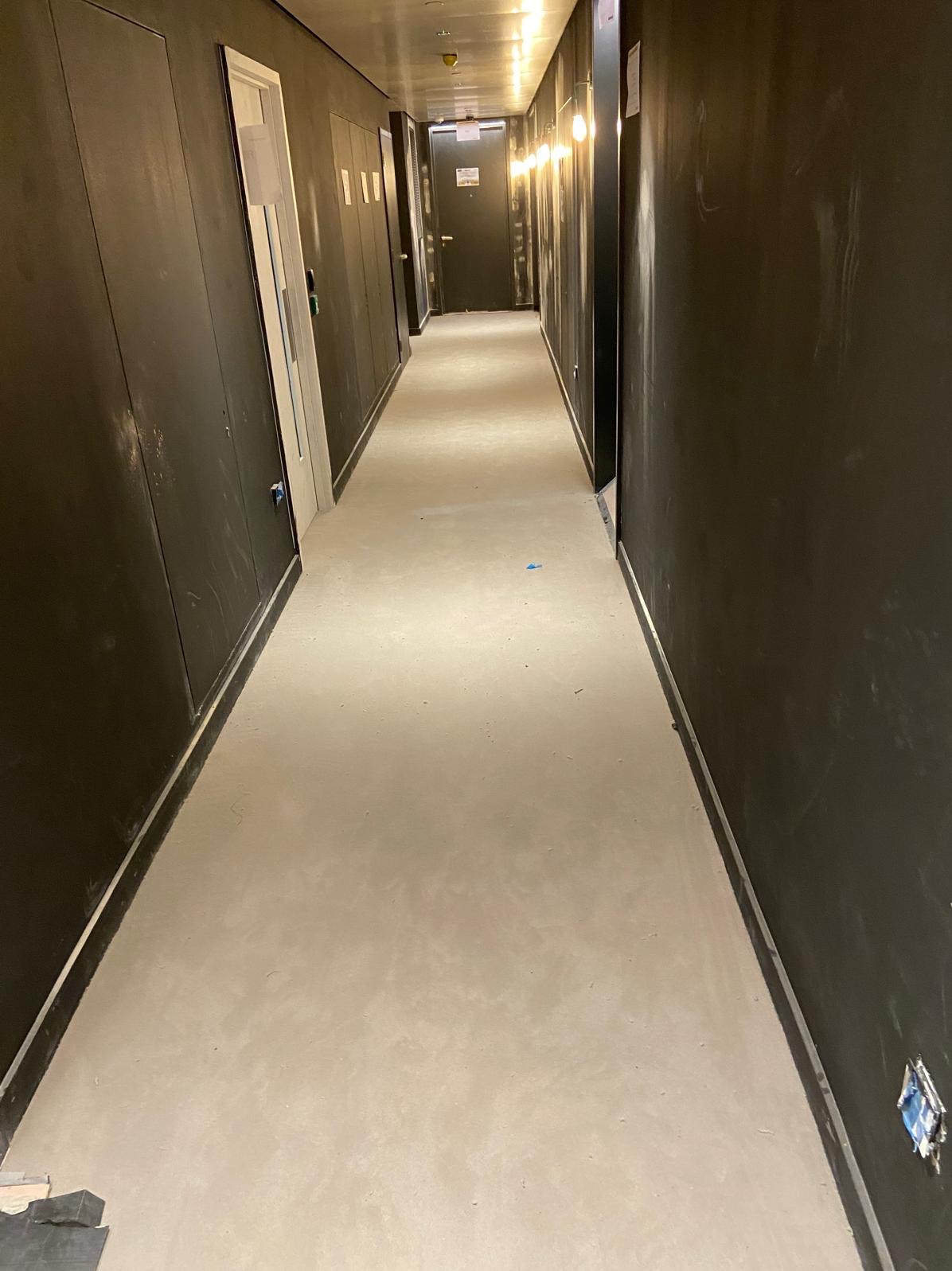 Długi korytarz z szarą wylewką samopoziomującą na podłodze i ciemnymi ścianami. Widoczne drzwi i oświetlenie sufitowe. W trakcie prac wykończeniowych.