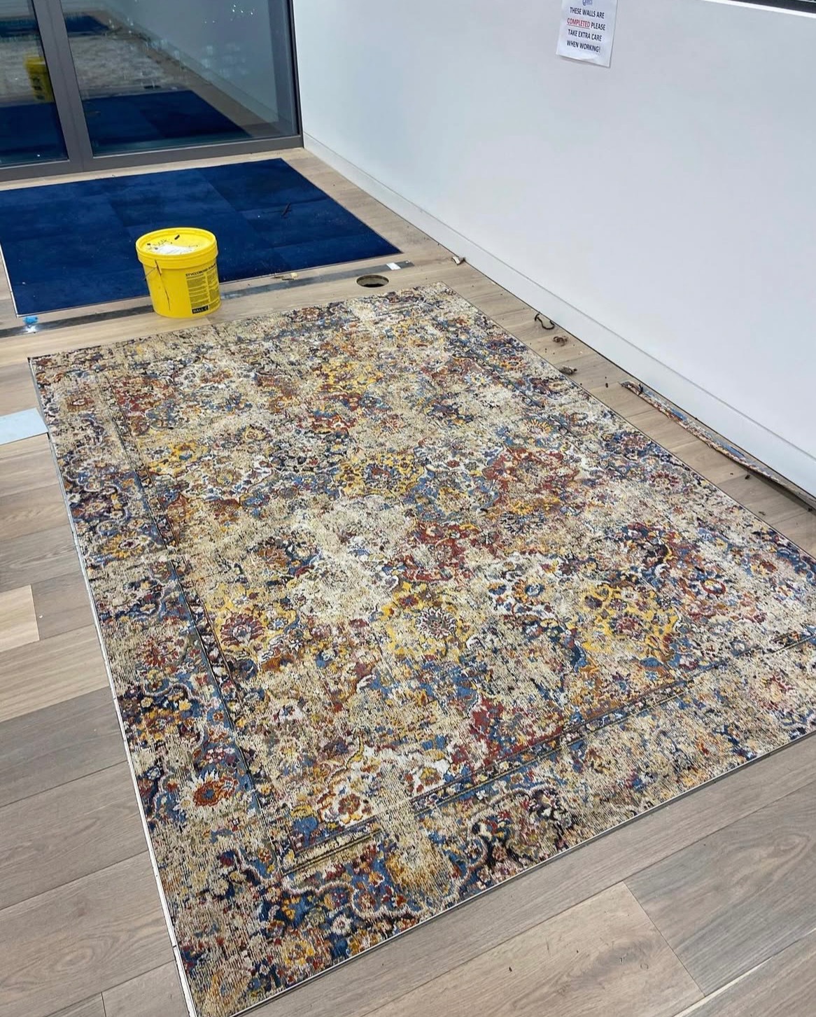 Fragment układania wzorzystego dywanu na drewnianej podłodze w jasnym wnętrzu. Żółte wiadro obok. Widoczne ślady po panelach podłogowych.