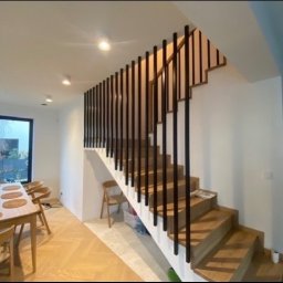 Prestiz Loft - Drewniane schody z czarnymi, pionowymi balustradami w nowoczesnym wnętrzu domu, widok na jadalnię z drewnianym stołem i krzesłami, parkiet w jodełkę.