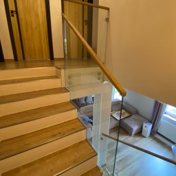 Prestiz Loft - Nowoczesne schody drewniane z balustradą szklaną i drewnianą poręczą. Widok z góry na salon z sofą i oknem. Estetyczne wykończenie wnętrza.