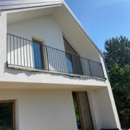 Nowoczesny dom z balkonem i czarną, metalową balustradą. Ujęcie z dołu ukazuje architekturę budynku i detale wykończenia.