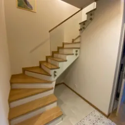 Drewniane schody z przeszkloną balustradą, mocowaną na ozdobnych uchwytach, w jasnym wnętrzu z obrazem na ścianie i wzorzystą podłogą.