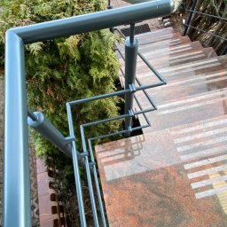 Prestiz Loft - Szare, metalowe poręcze schodowe z geometrycznym wzorem, widok z góry na marmurowe stopnie z białymi paskami, obok zielony krzew.