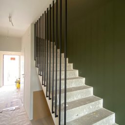 Prestiz Loft - Nowoczesne schody betonowe z czarną, minimalistyczną balustradą prętową. Widok na klatkę schodową w trakcie wykończenia, z zieloną ścianą w tle.
