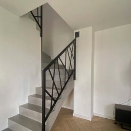 Prestiz Loft - Nowoczesne schody z szarą betonową konstrukcją i czarną, geometryczną poręczą. Parkiet w jodełkę na podłodze. Minimalistyczny design w jasnym wnętrzu.
