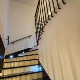 Prestiz Loft - Nowoczesne schody z czarnymi stopniami i podświetleniem LED, czarna metalowa balustrada z pionowymi prętami, drewniana poręcz na ścianie. Ujęcie z dołu.