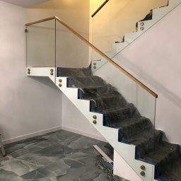 Prestiz Loft - Szklana balustrada z drewnianą poręczą na białych schodach, zabezpieczonych folią. Widoczne złote mocowania. Podłoga z szarego marmuru.