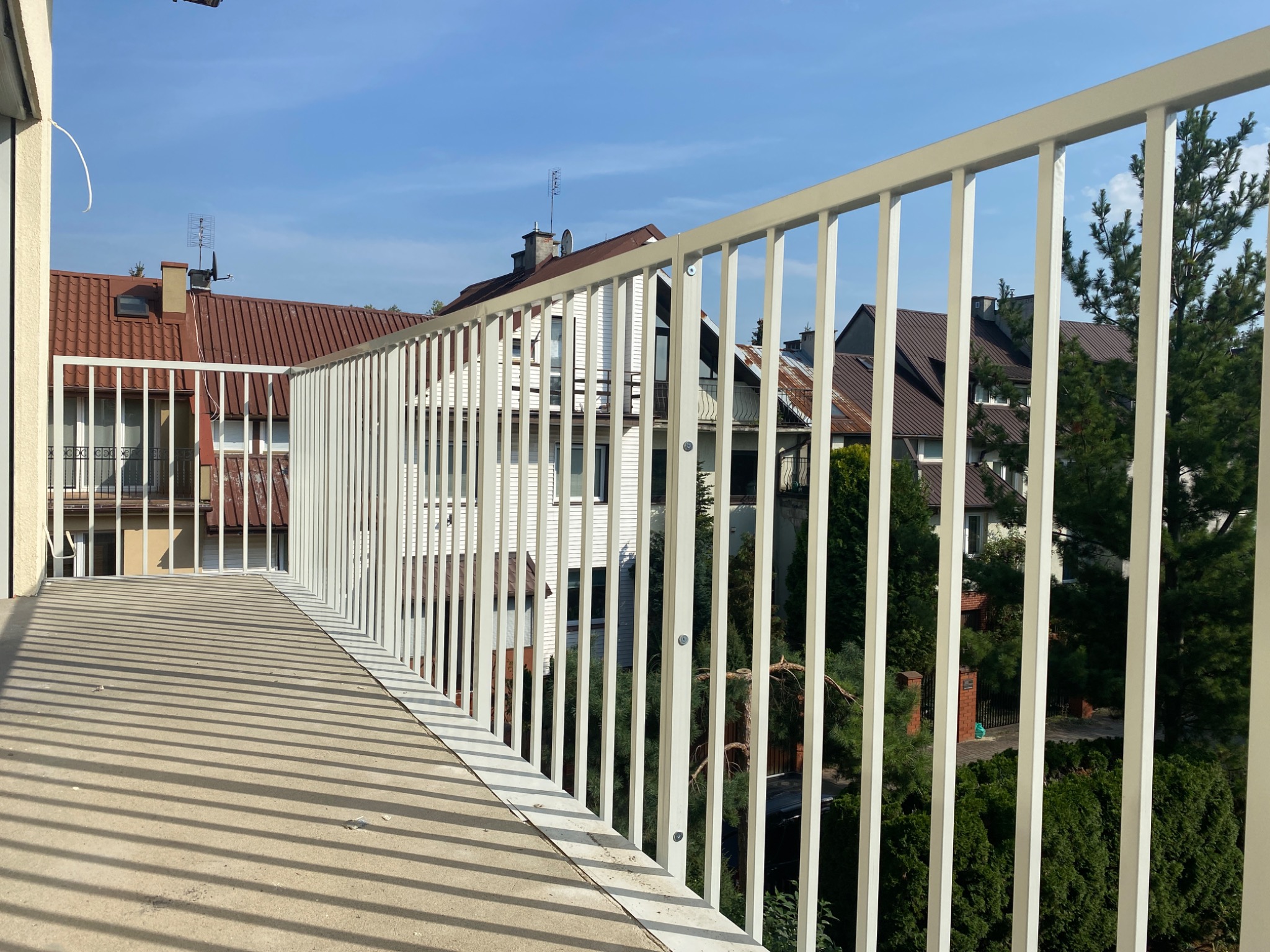 Biała, metalowa balustrada balkonowa z widokiem na zabudowę jednorodzinną i zieleń w słoneczny dzień, rzucająca cienie na betonową podłogę balkonu.