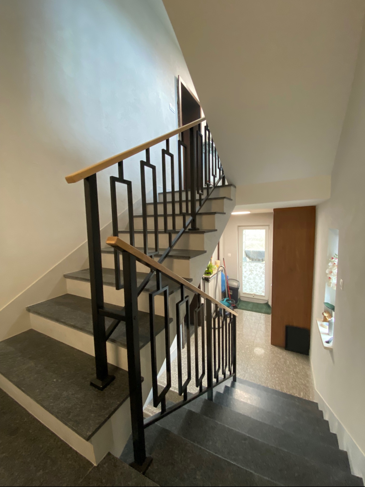 Wewnętrzne schody z ciemnego granitu z czarną metalową balustradą o geometrycznym wzorze i drewnianym poręczą, widok z dołu.