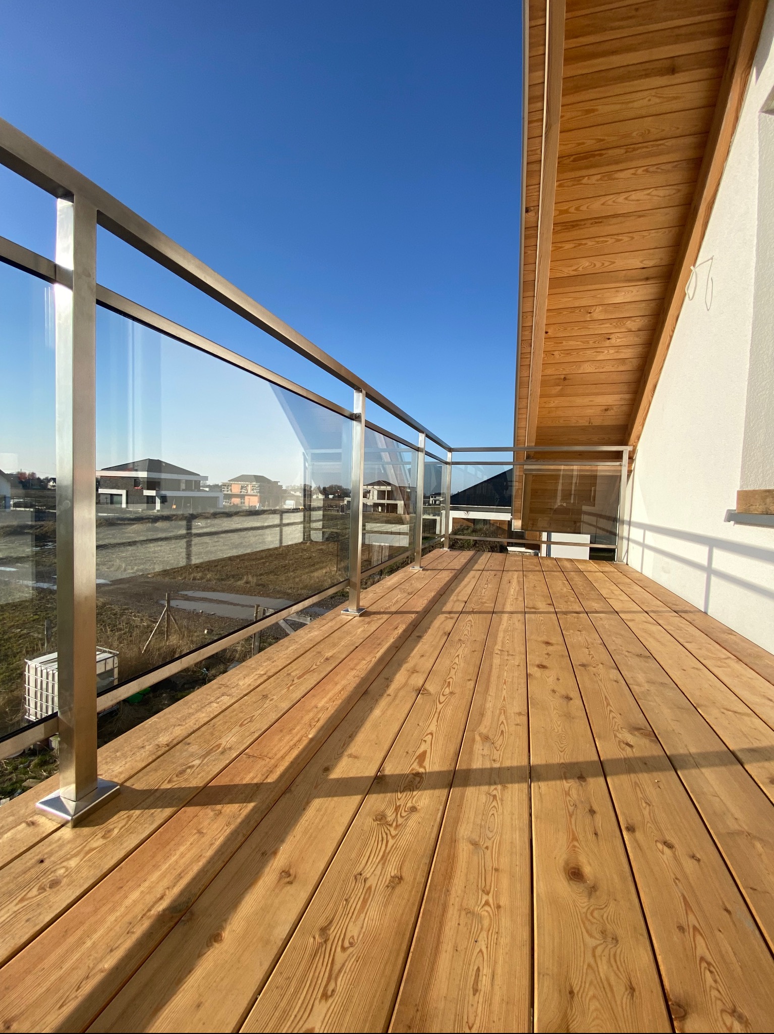 Balkon z drewnianą podłogą, balustradą ze stali nierdzewnej i szklanymi panelami, widok na nowe domy w tle, słoneczny dzień.