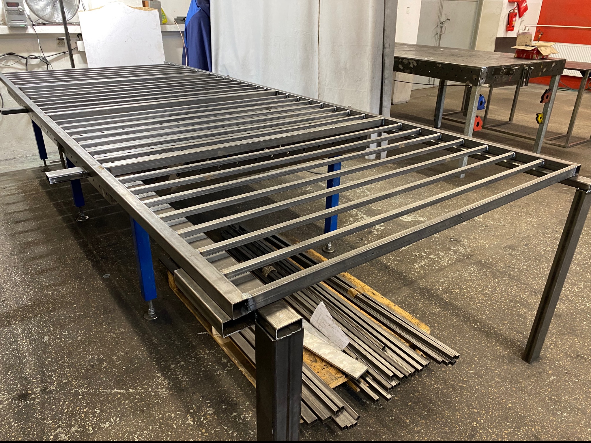 Konstrukcja metalowa w warsztacie w Warszawie, prawdopodobnie element balustrady lub bramy, spawana z profili kwadratowych, leżąca na stole roboczym, obok stos profili. Widoczne ślady obróbki.