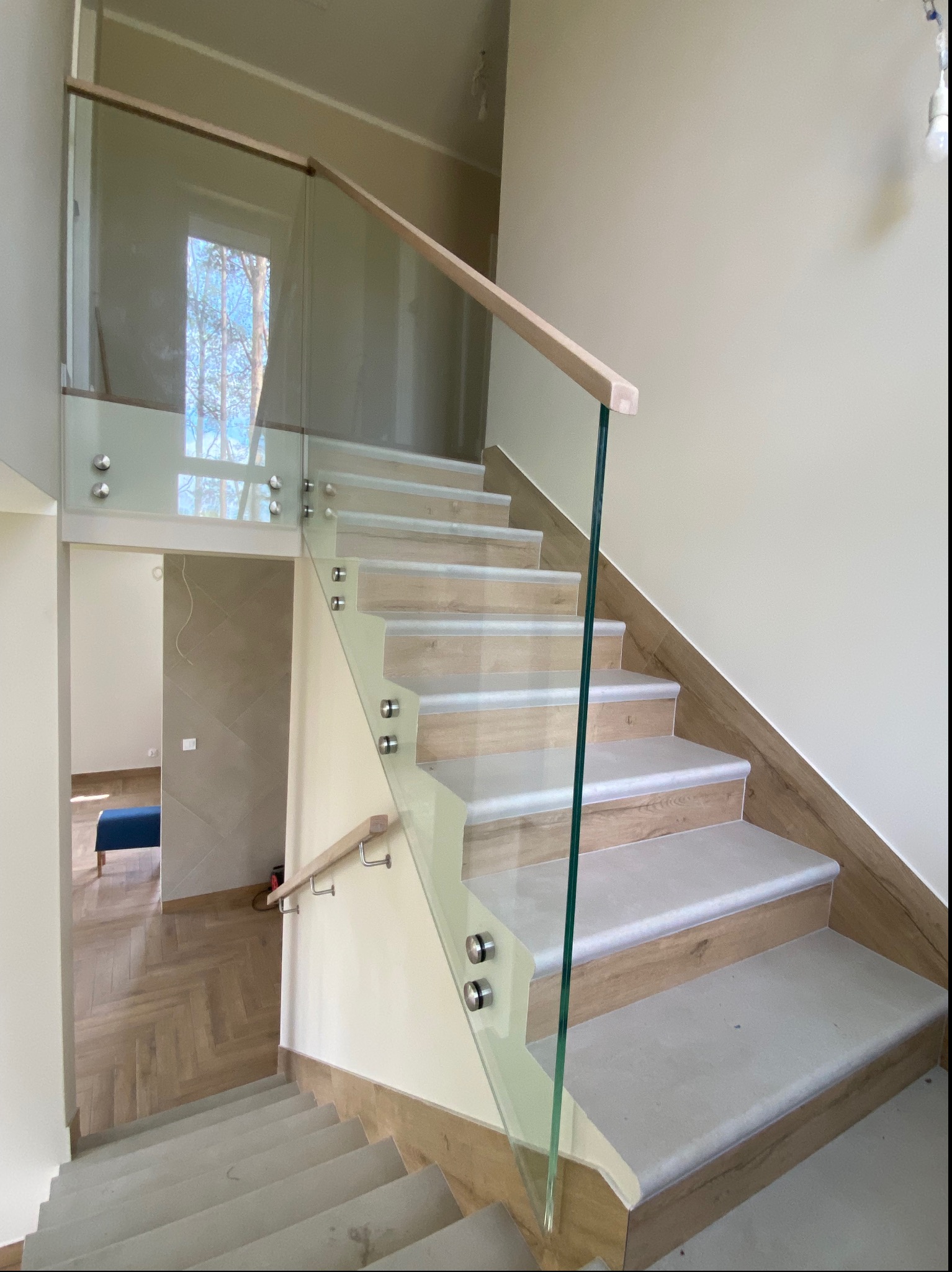 Nowoczesne schody z drewnianymi stopniami i szklaną balustradą mocowaną punktowo; widok na parkiet w jodełkę i fragment pomieszczenia z niebieskim siedziskiem.