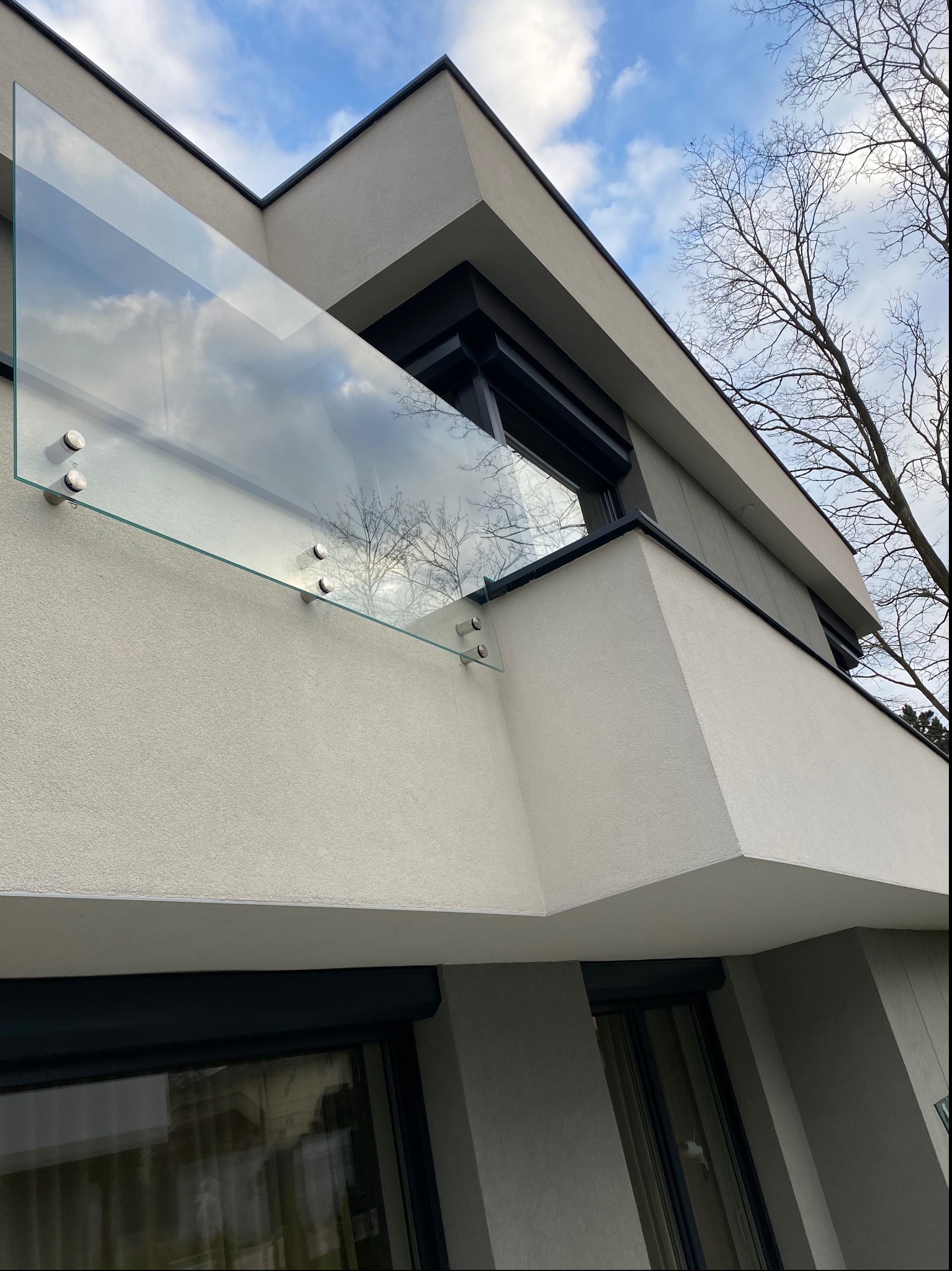 Nowoczesna szklana balustrada balkonowa zamocowana punktowo do elewacji budynku o minimalistycznej architekturze, na tle błękitnego nieba z chmurami i gałęzi drzew bez liści.