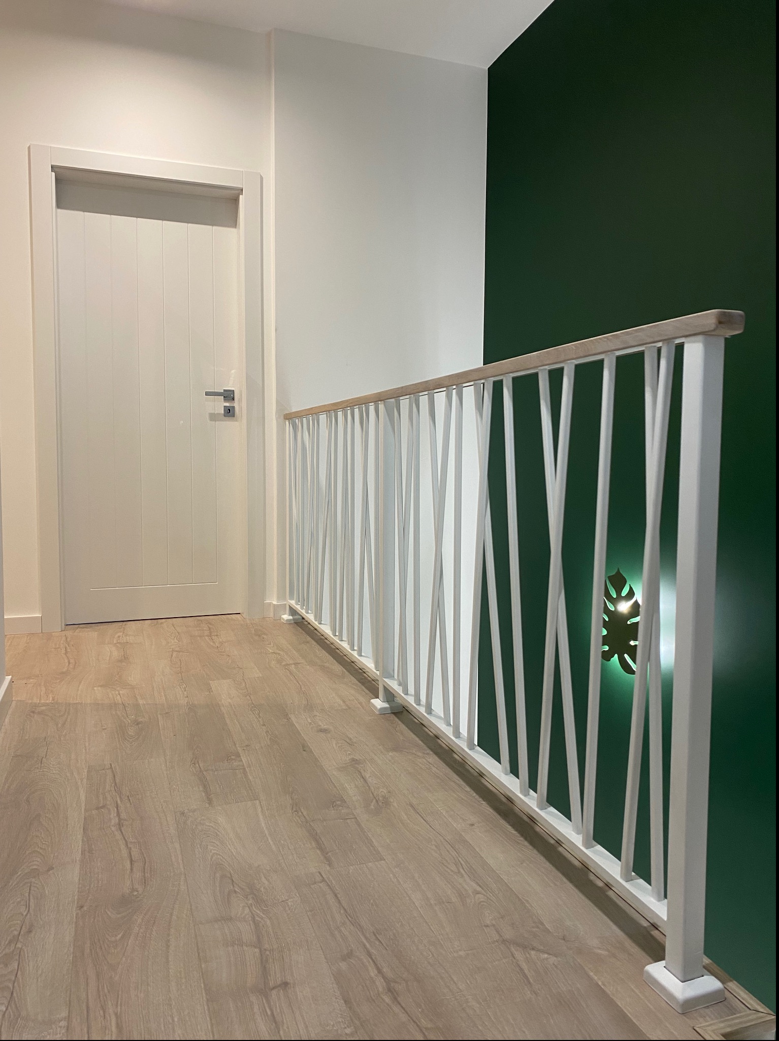 Nowoczesna balustrada wewnętrzna z białymi, ukośnymi szczeblami i drewnianym poręczą na tle zielonej ściany z podświetlanym motywem liścia, widoczna na korytarzu z panelami podłogowymi i białymi...