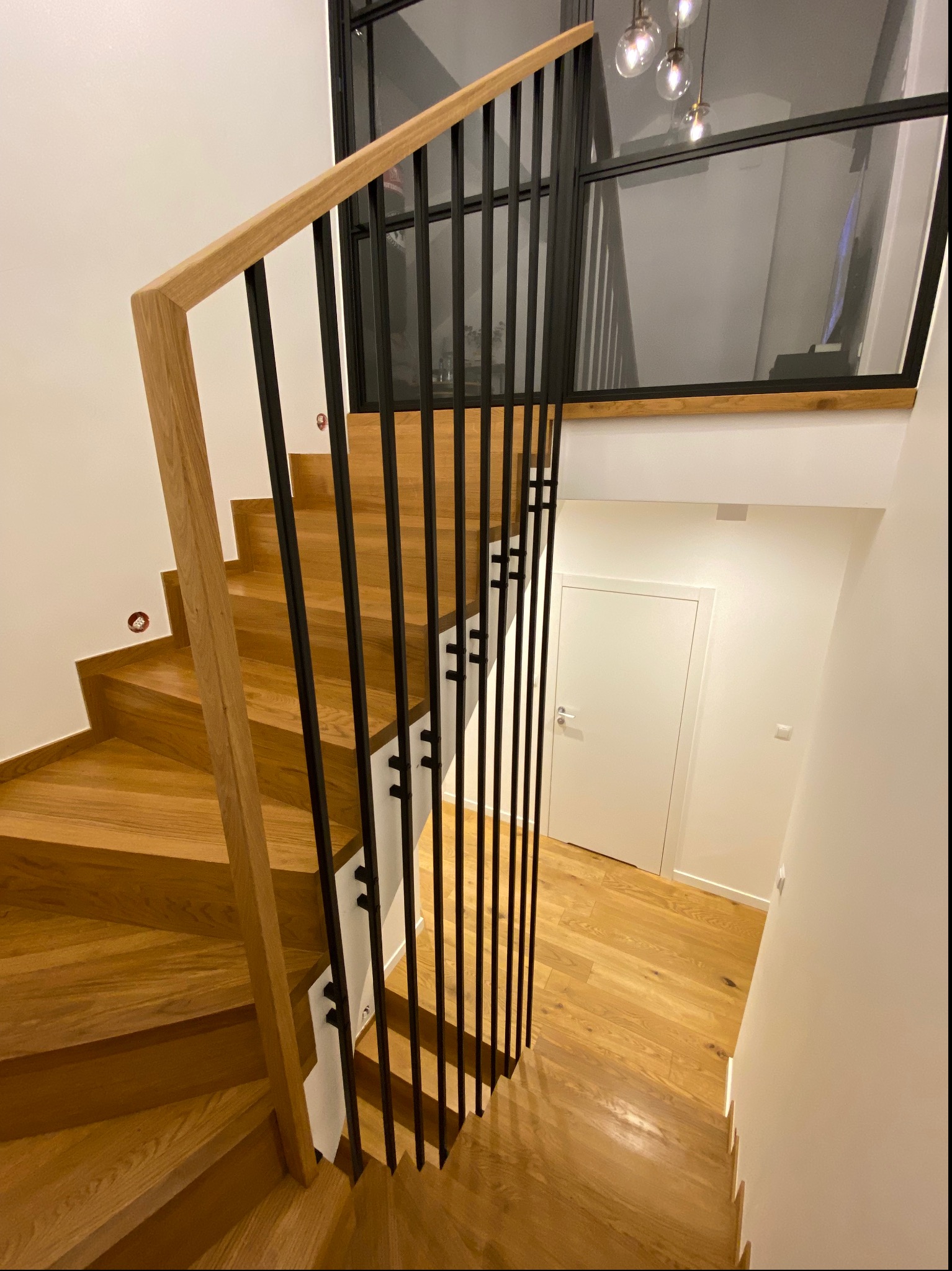 Drewniane schody z czarną, metalową balustradą z pionowych prętów i drewnianą poręczą, widok z góry, białe ściany i drzwi w tle.