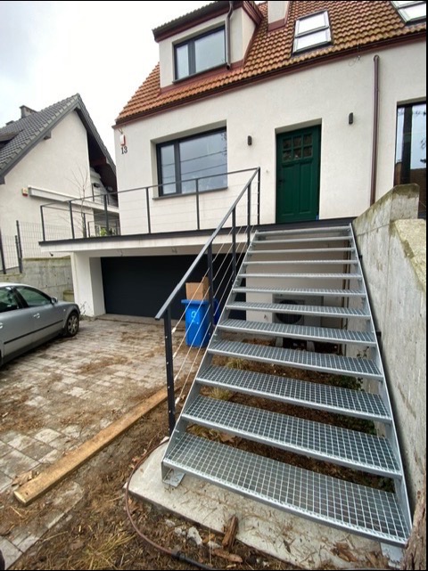 Zewnętrzne schody metalowe z kraty pomostowej z balustradą z linek stalowych prowadzące do domu z zielonymi drzwiami i balkonem z czarną balustradą.
