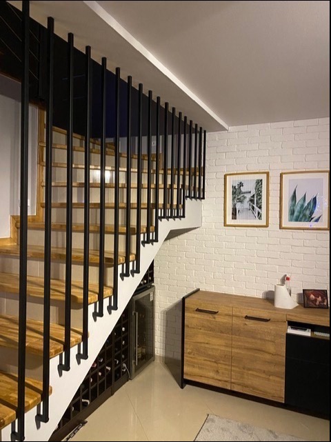 Nowoczesne schody z drewnianymi stopniami i czarną, pionową balustradą, w tle ściana z białej cegły i komoda w odcieniach brązu i czerni, pod schodami wbudowana chłodziarka na wino.