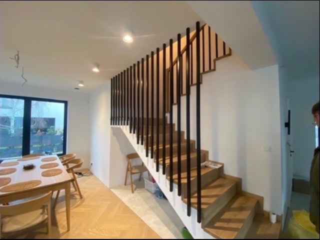 Drewniane schody z czarnymi, pionowymi balustradami w nowoczesnym wnętrzu domu, widok na jadalnię z drewnianym stołem i krzesłami, parkiet w jodełkę.