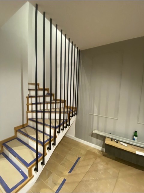 Wnętrze domu z drewnianymi schodami z białymi podstopnicami zabezpieczonymi taśmą malarską i pionową, czarną balustradą z okrągłych prętów.