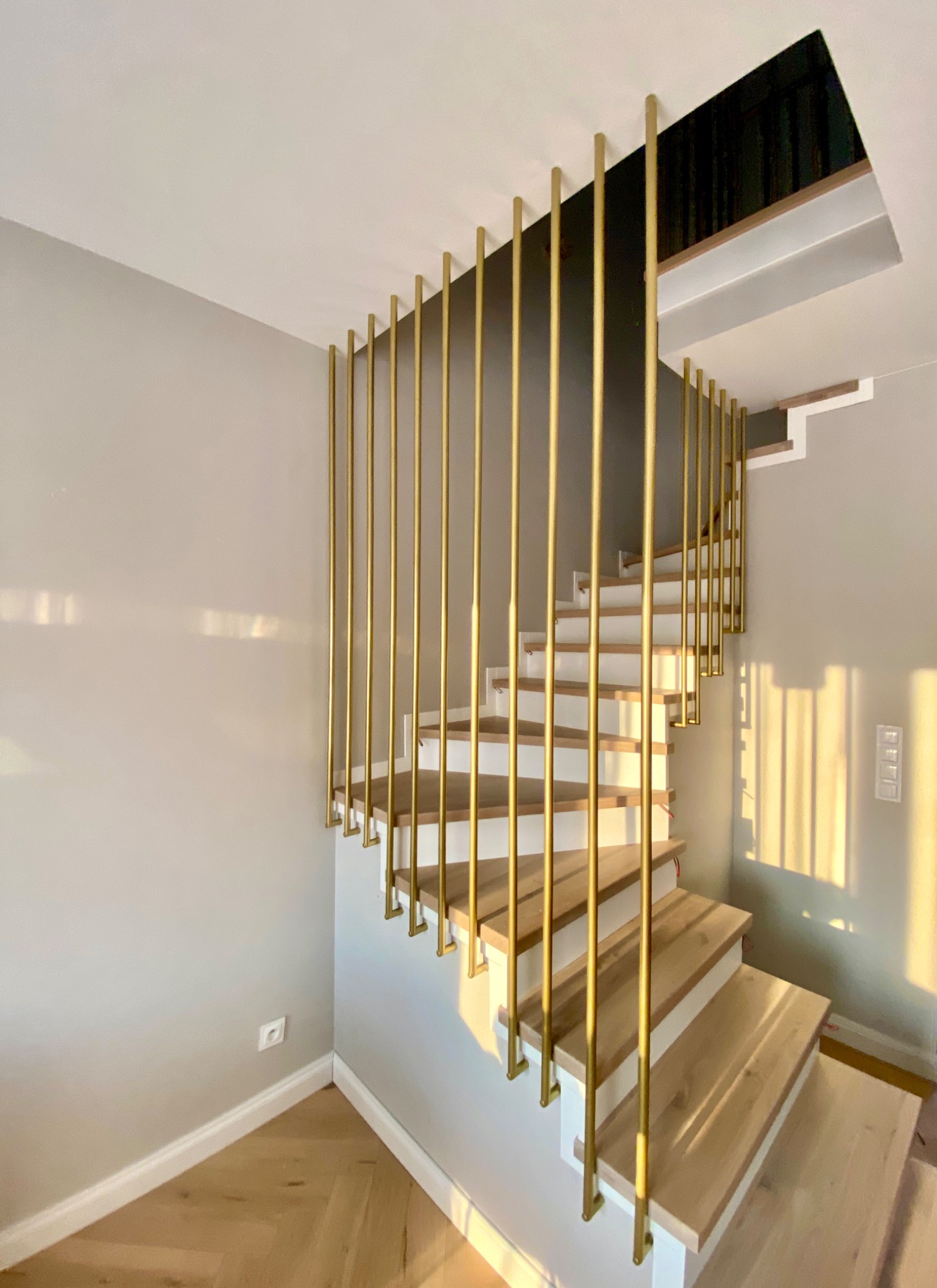 Nowoczesne schody wewnętrzne z drewnianymi stopniami i złotymi, pionowymi balustradami z rurek, widok z dołu na jasne wnętrze z parkietem i szarą ścianą.