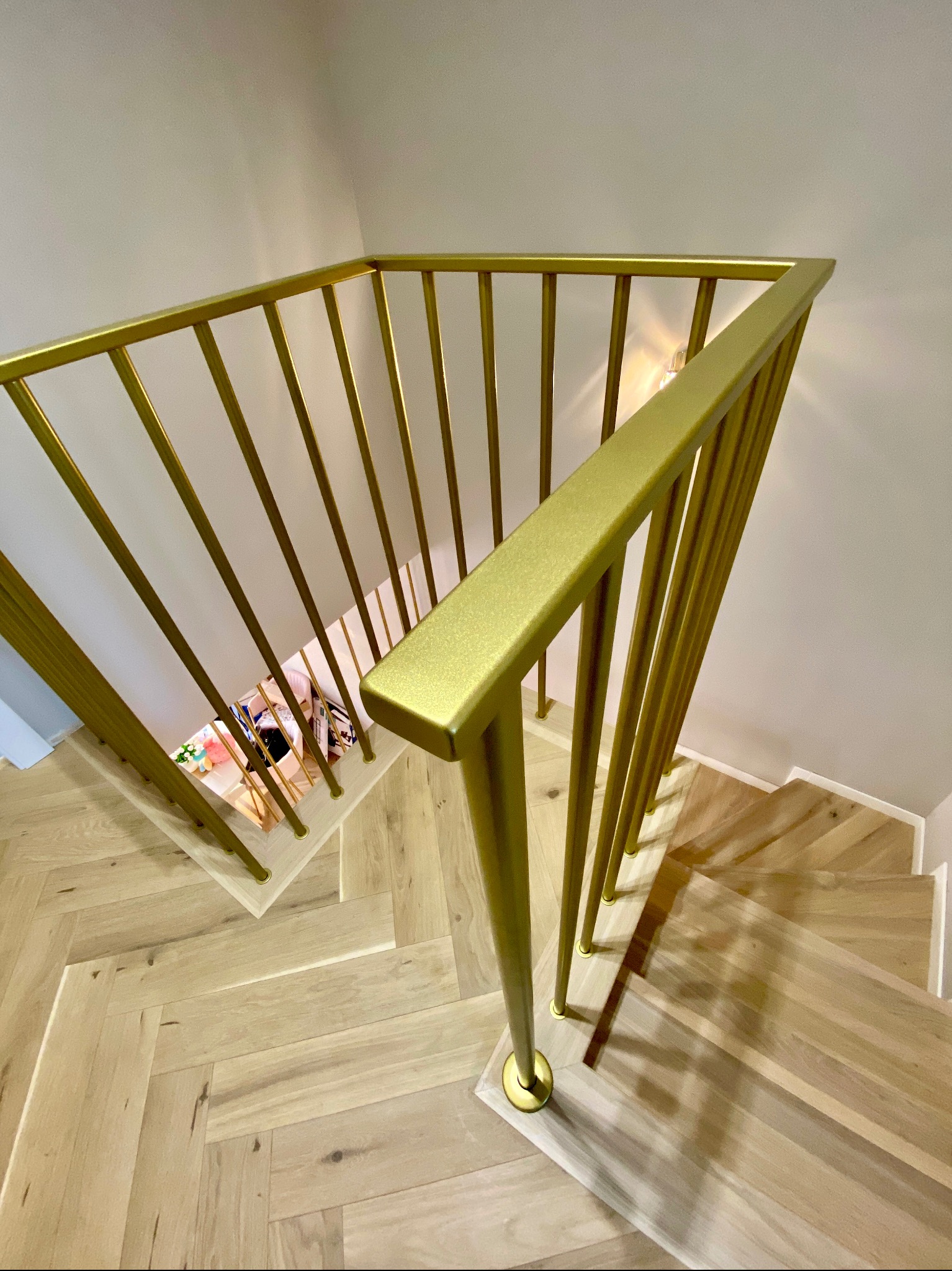Widok z góry na złote, metalowe poręcze schodowe z pionowymi prętami, zamontowane na drewnianych schodach ułożonych w jodełkę; w tle widoczne dziecięce zabawki.