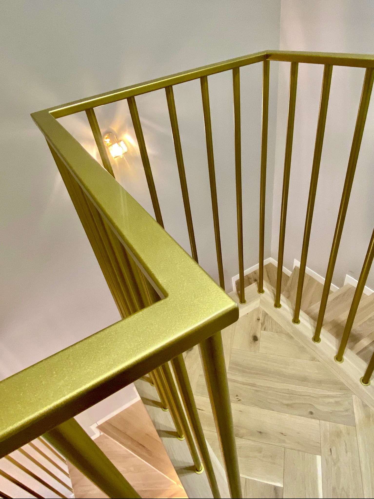 Złota balustrada schodowa z pionowymi prętami, widok z góry na drewniane schody ułożone w jodełkę, oświetlenie punktowe w tle.