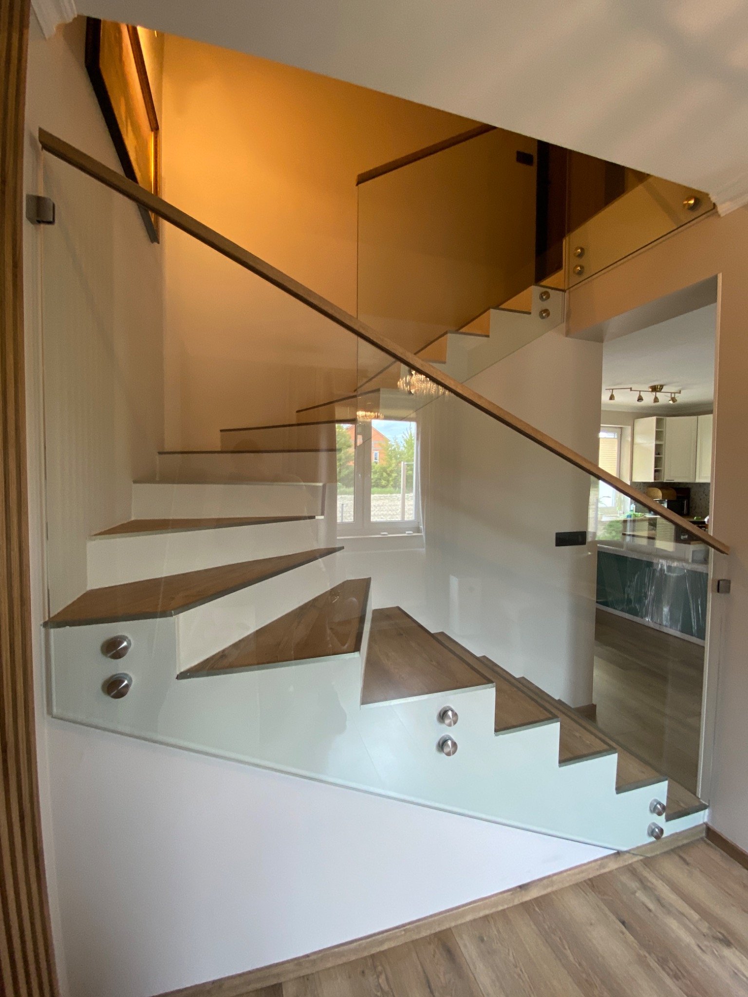 Nowoczesne schody z drewnianymi stopniami i szklaną balustradą mocowaną punktowo. Widok z dołu, z widoczną kuchnią w tle. Jasne wnętrze, minimalistyczny design.