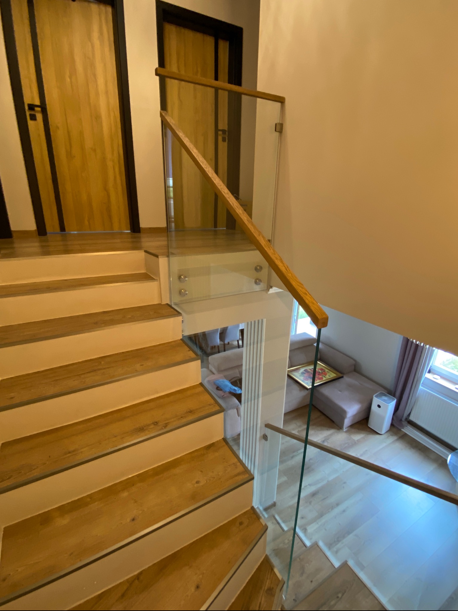 Nowoczesne schody drewniane z balustradą szklaną i drewnianą poręczą. Widok z góry na salon z sofą i oknem. Estetyczne wykończenie wnętrza.