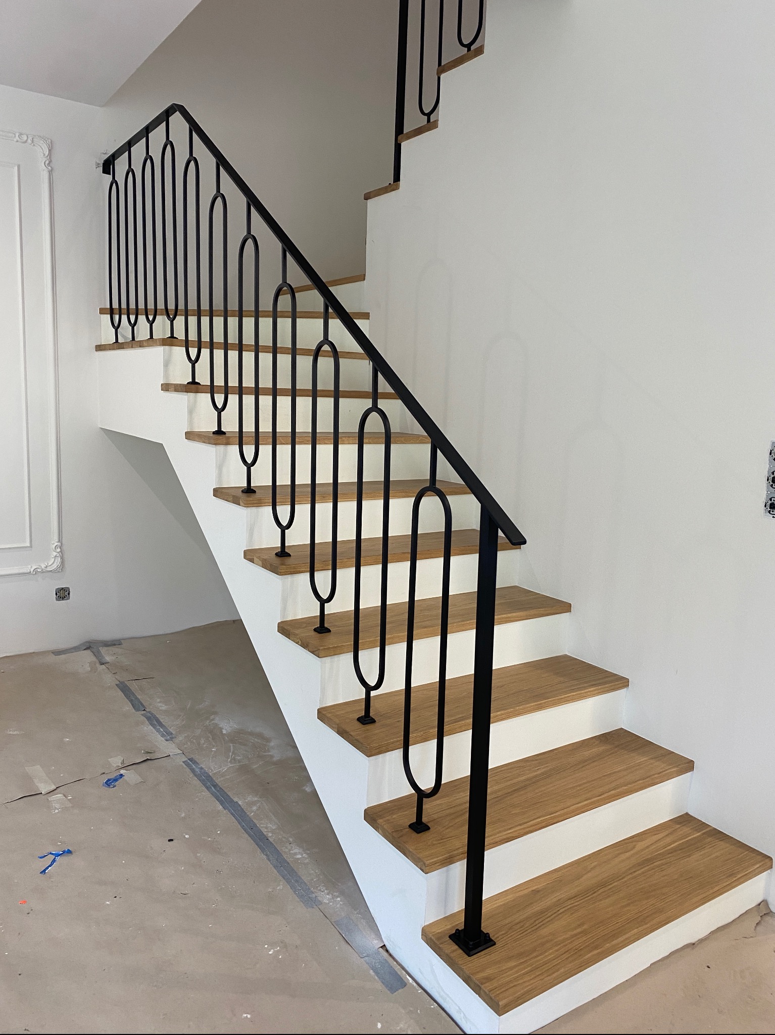 Nowoczesne schody z drewnianymi stopniami i czarną, metalową balustradą o unikalnym wzorze, w jasnym wnętrzu. Widoczne detale wykończeniowe i surowe podłoże.