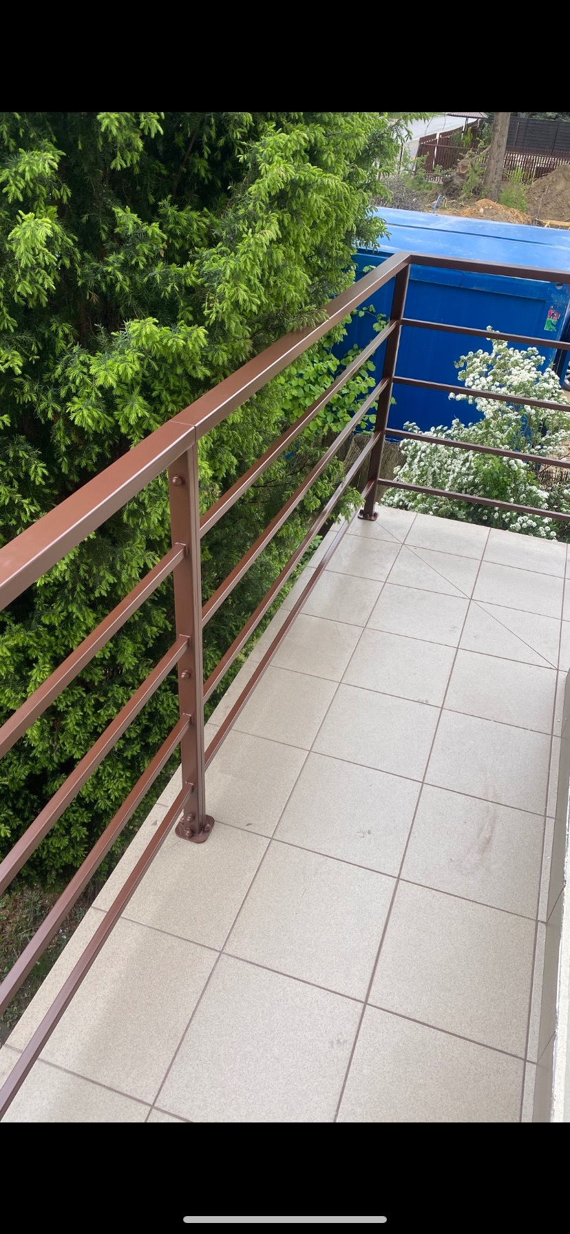 Balkon z brązową, metalową konstrukcją balustrady, widok z góry. W tle zieleń krzewów i błękitna plandeka. Płytki podłogowe w neutralnym odcieniu.