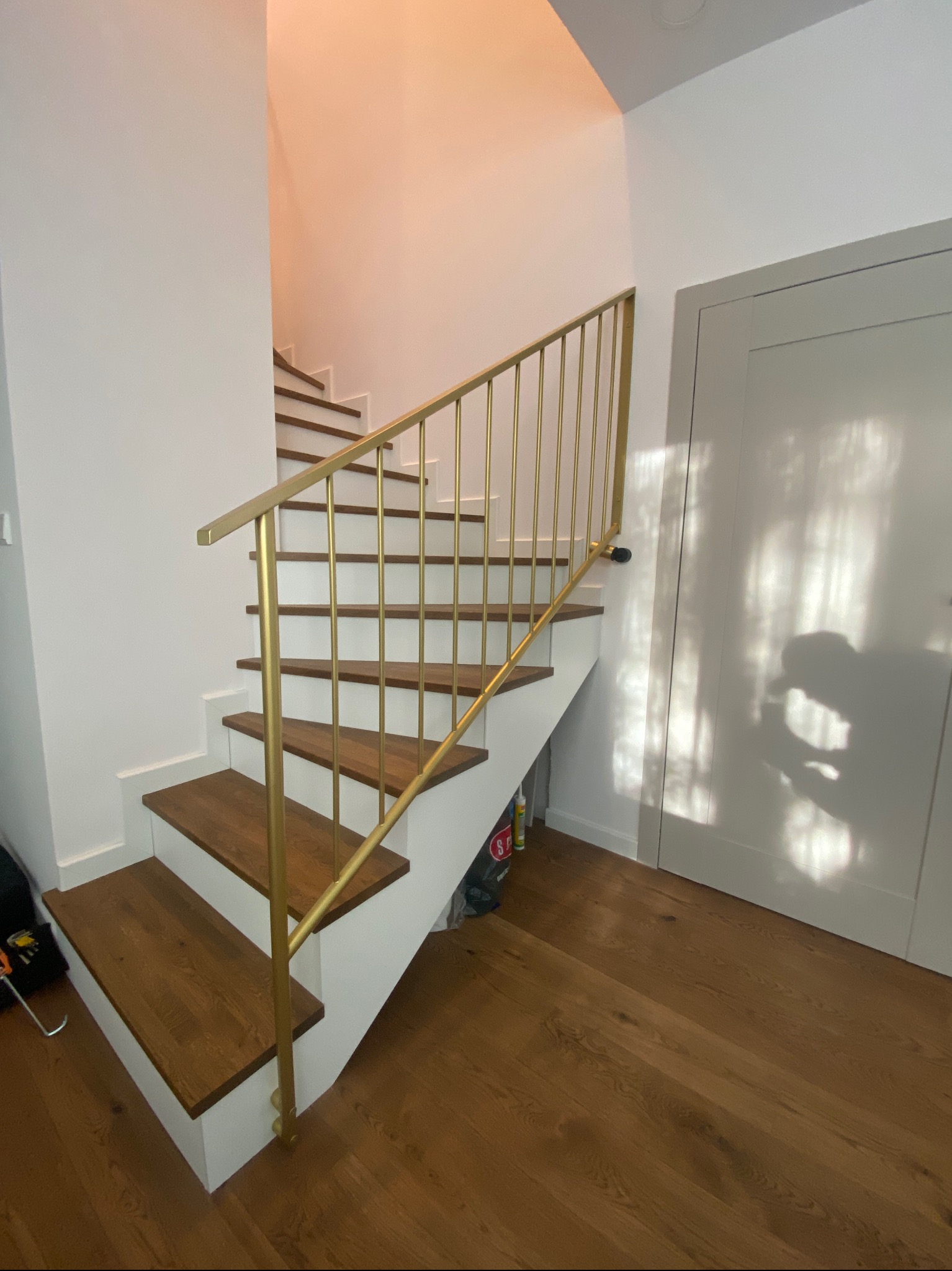 Schody z drewnianymi stopniami i złotą, metalową poręczą. Na ścianie cień osoby. Widok z dołu na konstrukcję schodów i minimalistyczną poręcz.