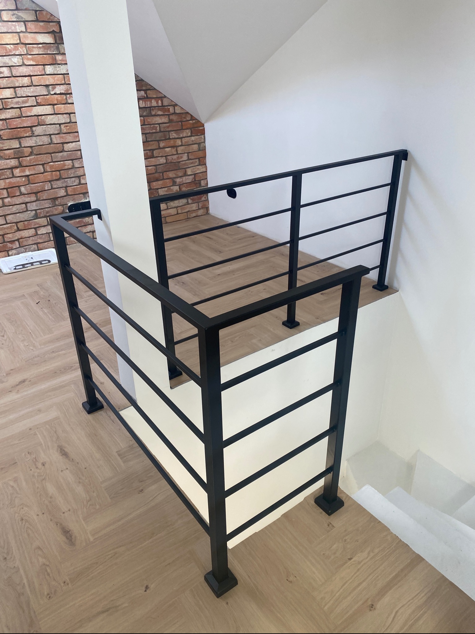 Nowoczesna, czarna, metalowa barierka schodowa z poziomymi szczeblami, zamontowana w jasnym wnętrzu z drewnianą podłogą i fragmentem ściany z czerwonej cegły.