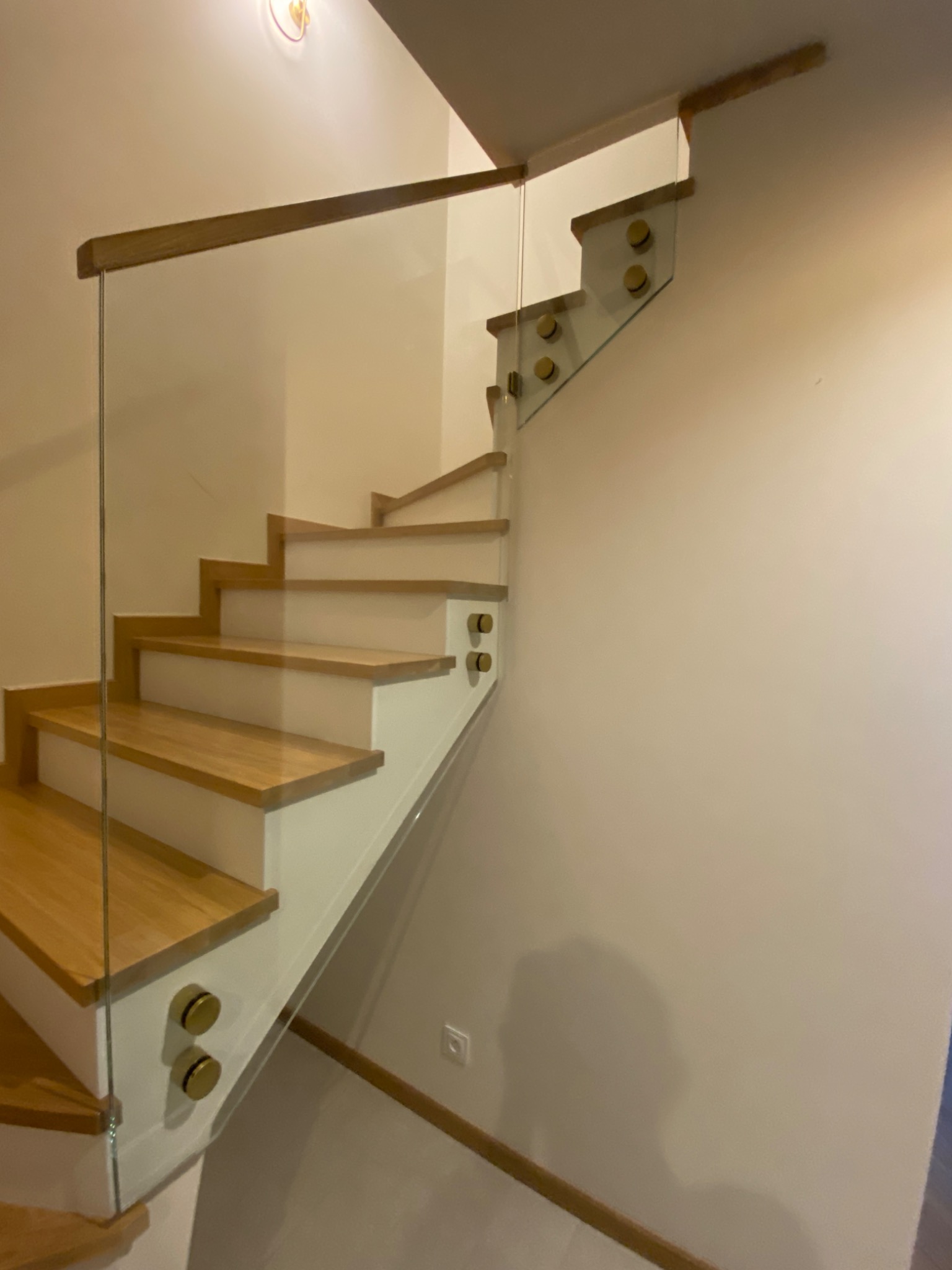 Nowoczesne schody drewniane z białymi podstopnicami i szklaną balustradą mocowaną złotymi uchwytami, widoczne z boku, oświetlone ciepłym światłem.