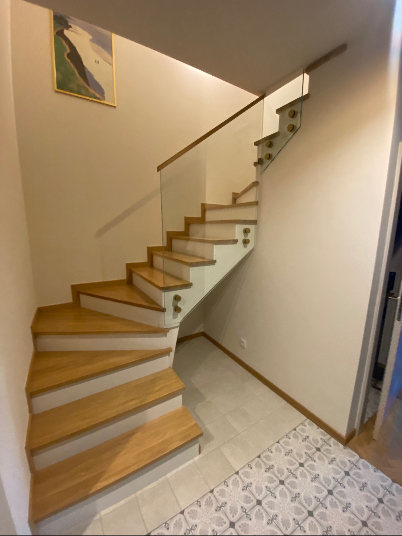 Drewniane schody z przeszkloną balustradą, mocowaną na ozdobnych uchwytach, w jasnym wnętrzu z obrazem na ścianie i wzorzystą podłogą.