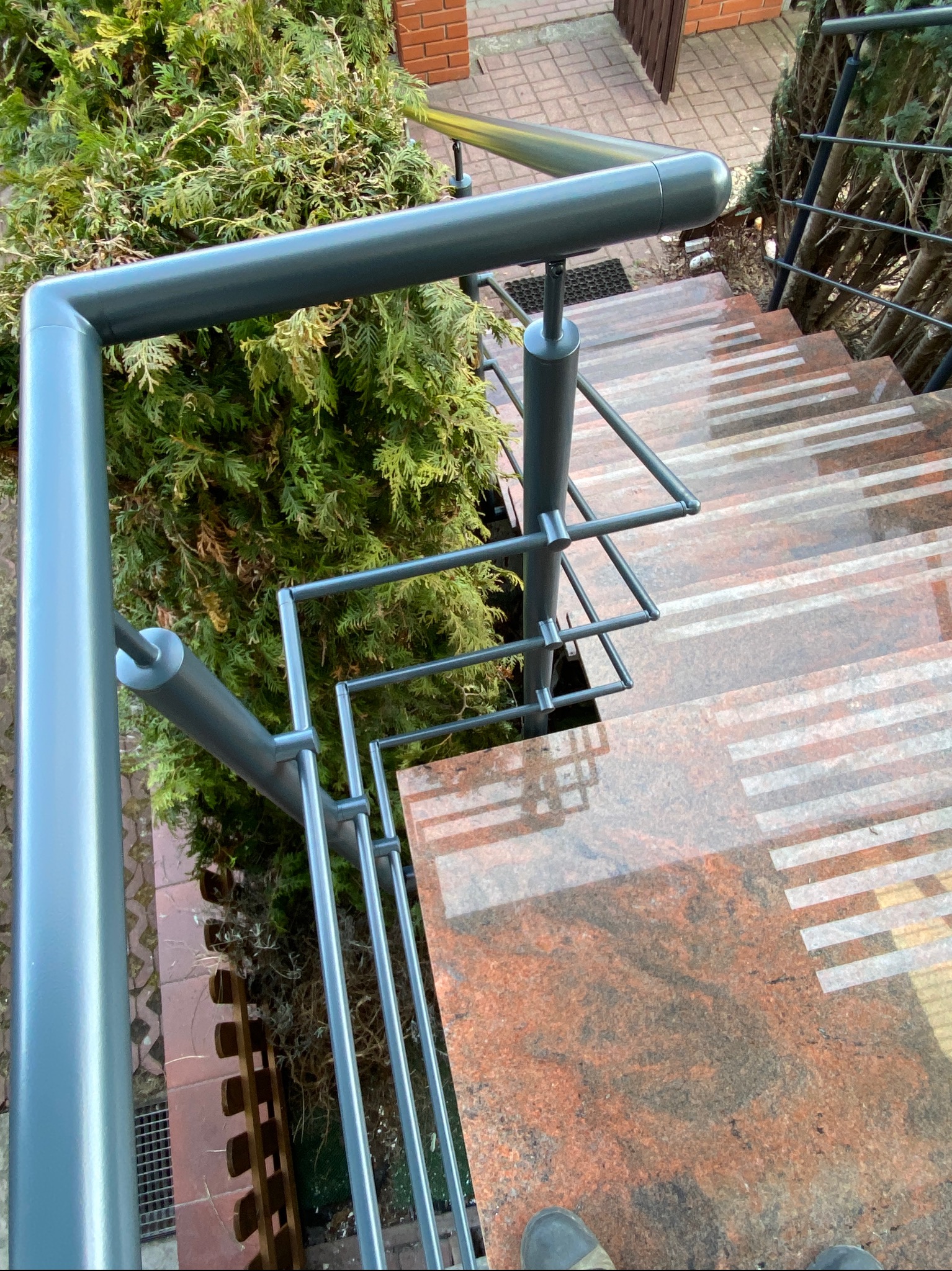 Szare, metalowe poręcze schodowe z geometrycznym wzorem, widok z góry na marmurowe stopnie z białymi paskami, obok zielony krzew.