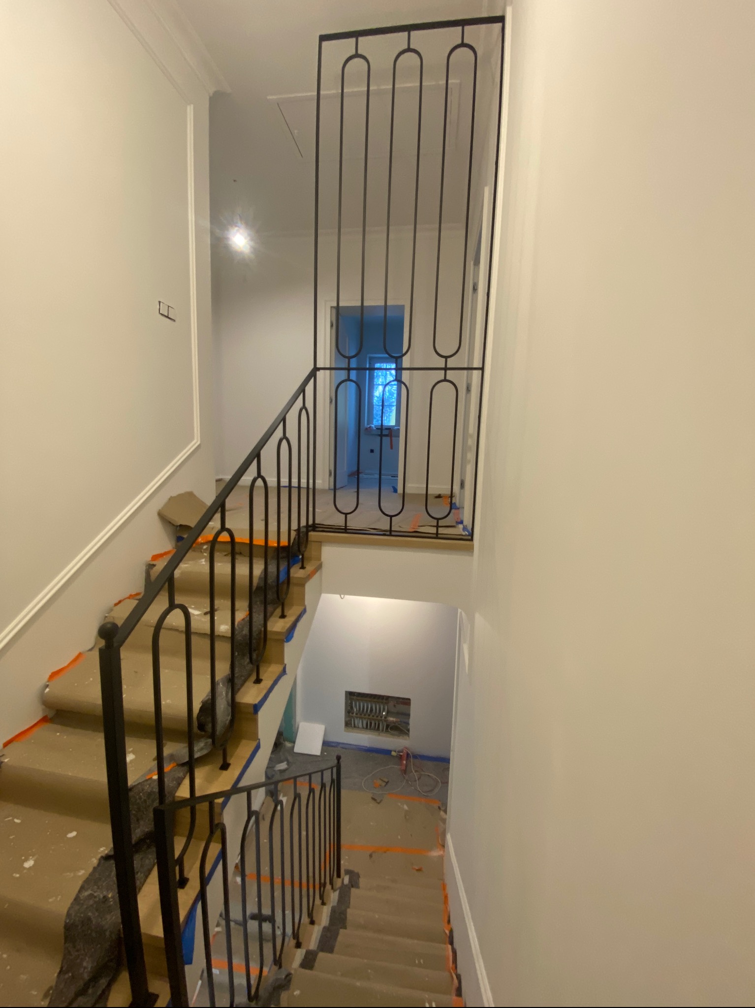 Nowoczesne, czarne poręcze schodowe z motywem zaokrąglonych prostokątów, widoczne z góry. Schody zabezpieczone folią malarską, wnętrze w trakcie wykończenia.