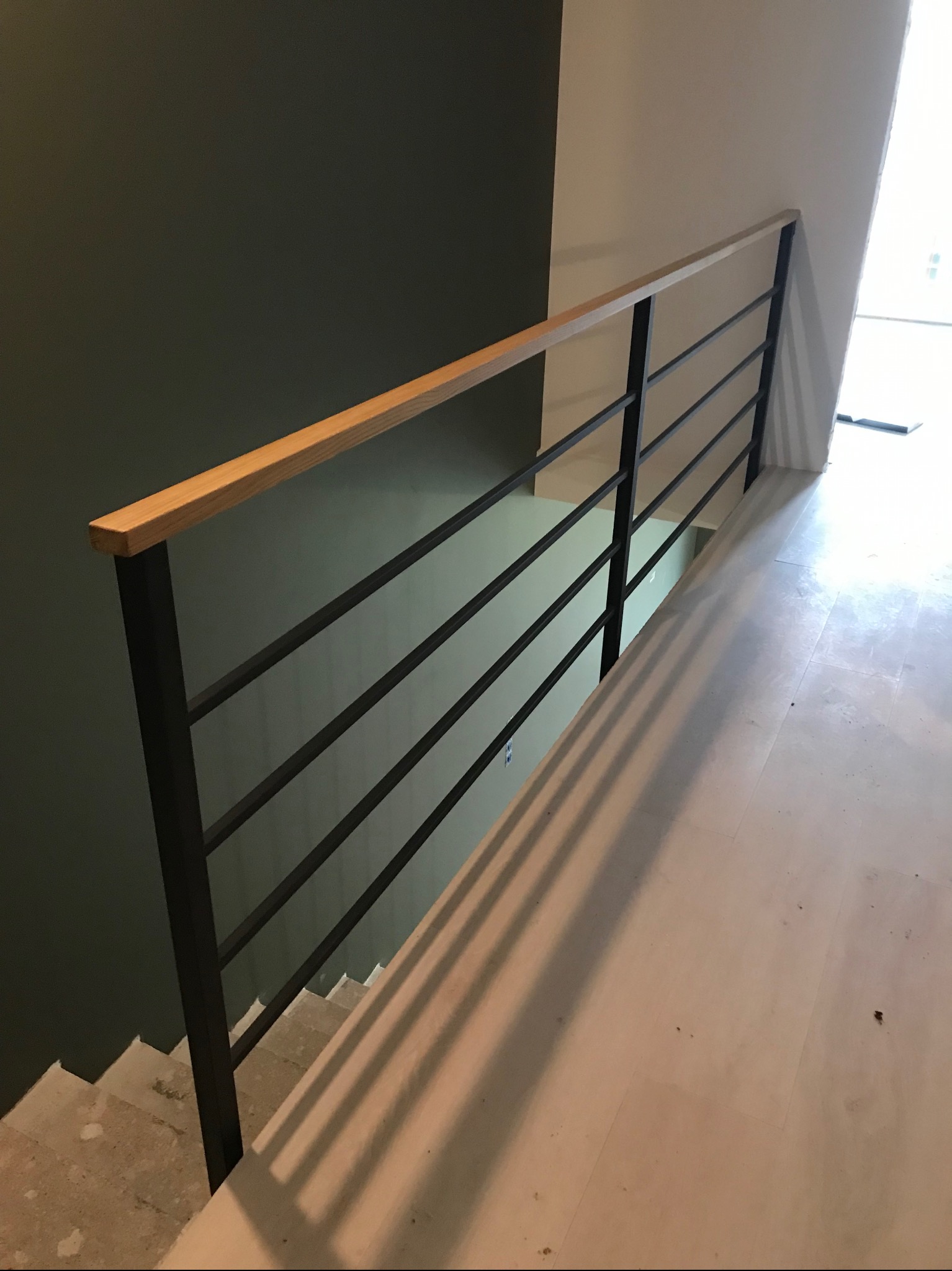 Minimalistyczna poręcz schodowa z czarnych, poziomych prętów i drewnianego uchwytu, zamontowana na tle ściany w dwóch odcieniach szarości, z widocznymi schodami.