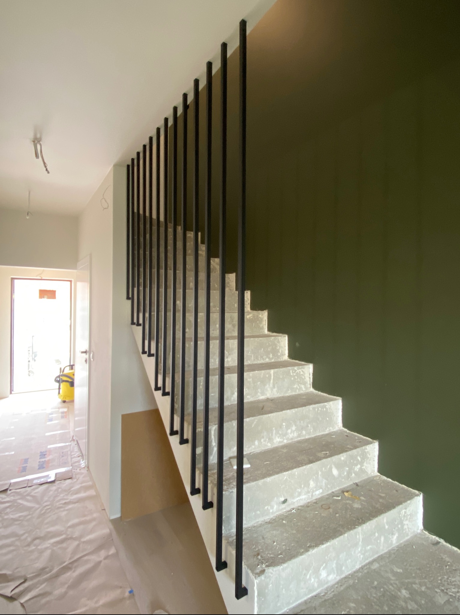 Nowoczesne schody betonowe z czarną, minimalistyczną balustradą prętową. Widok na klatkę schodową w trakcie wykończenia, z zieloną ścianą w tle.