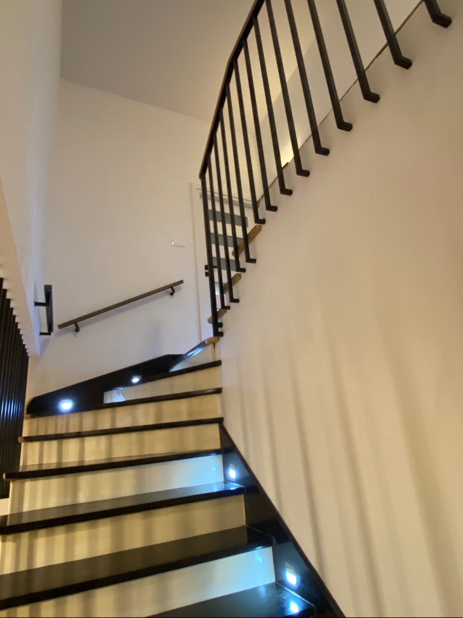 Nowoczesne schody z czarnymi stopniami i podświetleniem LED, czarna metalowa balustrada z pionowymi prętami, drewniana poręcz na ścianie. Ujęcie z dołu.