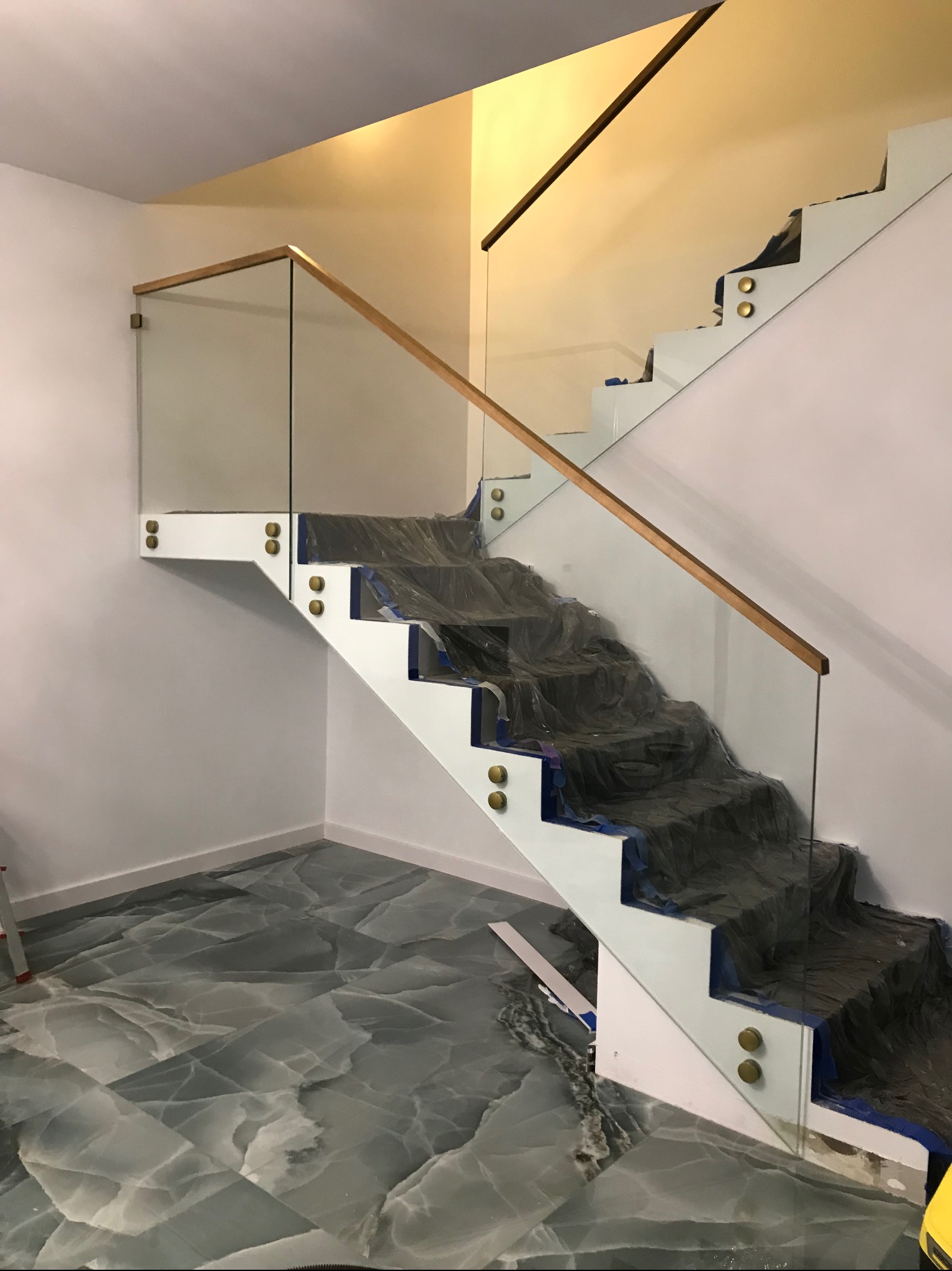 Szklana balustrada z drewnianą poręczą na białych schodach, zabezpieczonych folią. Widoczne złote mocowania. Podłoga z szarego marmuru.