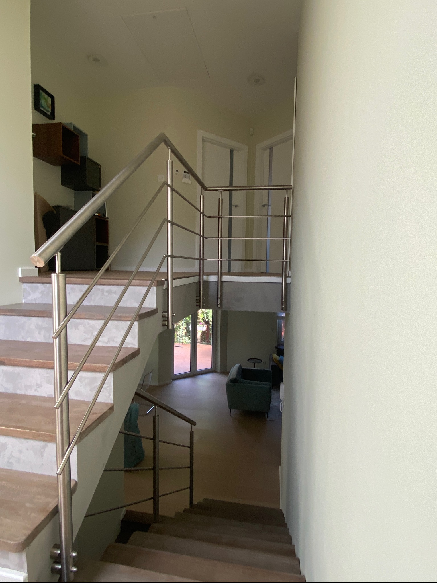 Wnętrze domu z drewnianymi schodami i nowoczesną, metalową balustradą. Widok na salon z antresoli. Jasne ściany i naturalne światło.