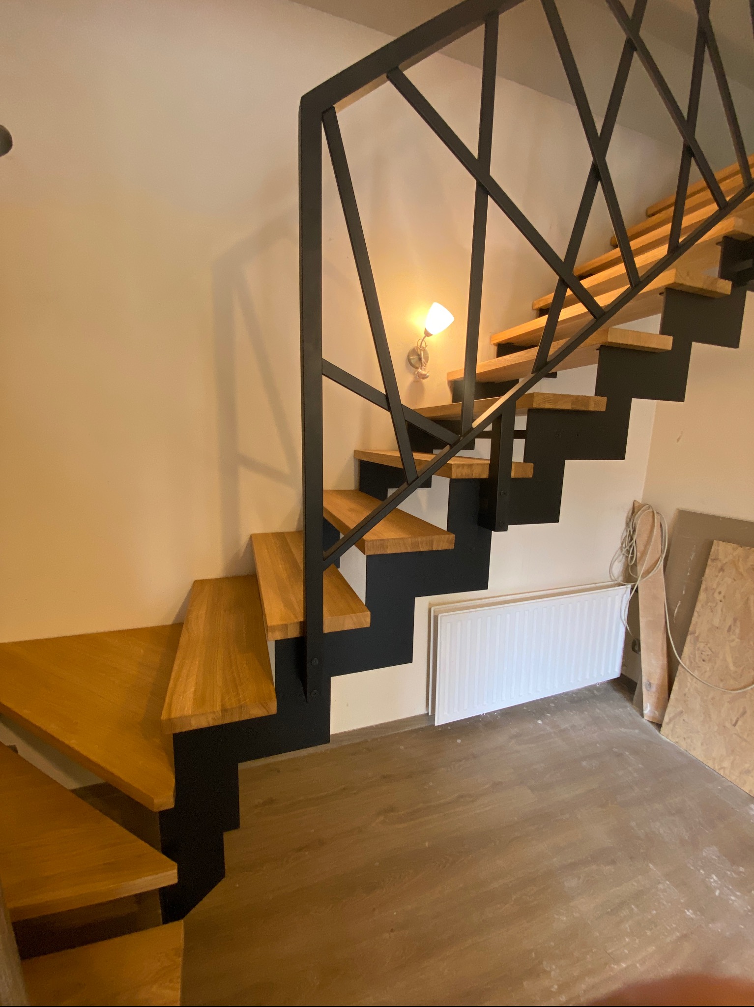 Nowoczesne schody z drewnianymi stopniami i czarną, geometryczną konstrukcją. Widoczny fragment ściany z kinkietem i elementy wykończeniowe w tle.