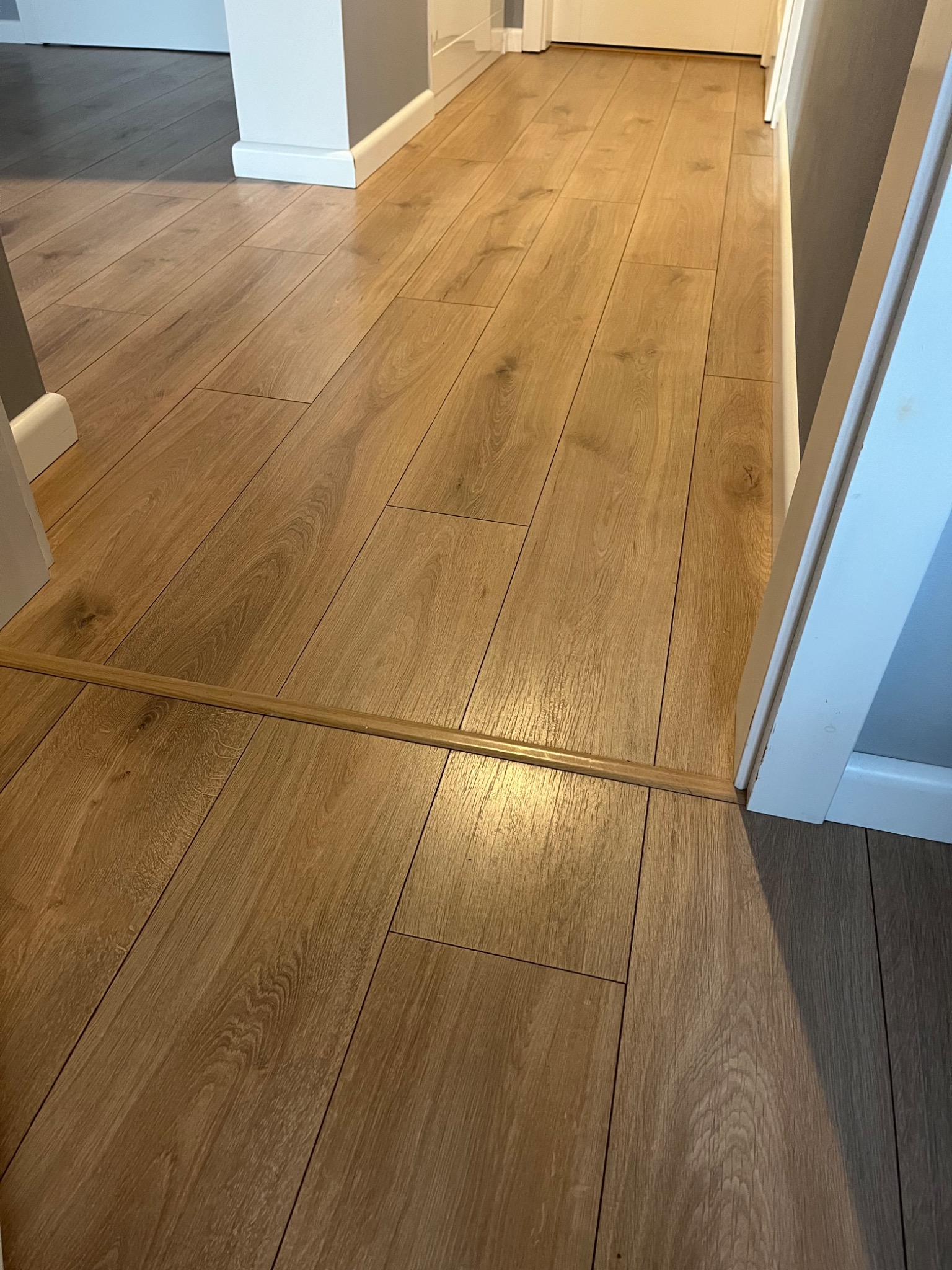 Jasne, drewnopodobne panele podłogowe z listwą progową łączącą dwa pomieszczenia, widoczne białe listwy przypodłogowe i fragment ościeżnicy drzwiowej.