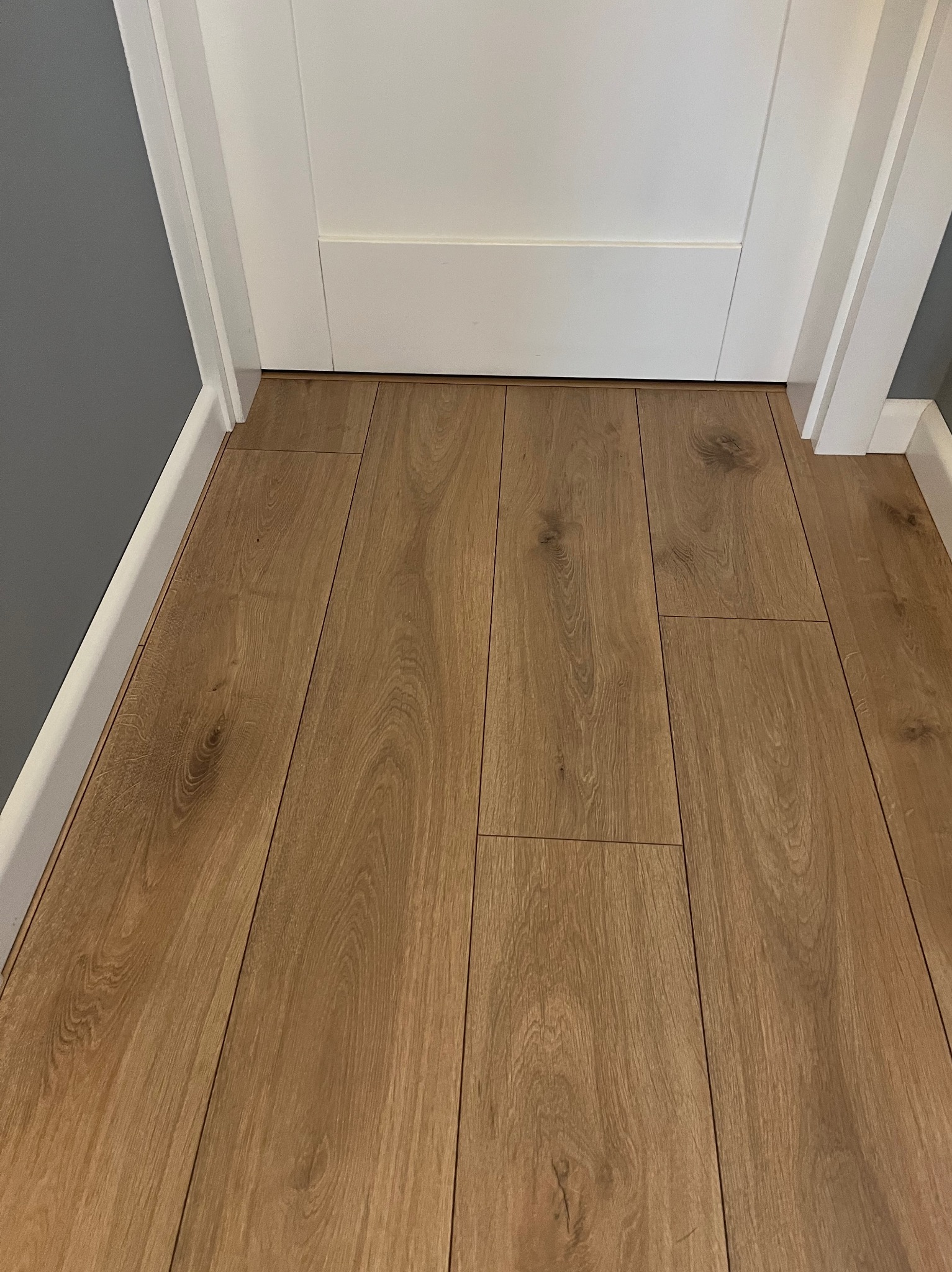 Ułożone panele podłogowe w odcieniach brązu, przylegające do białej futryny drzwiowej i szarej ściany.