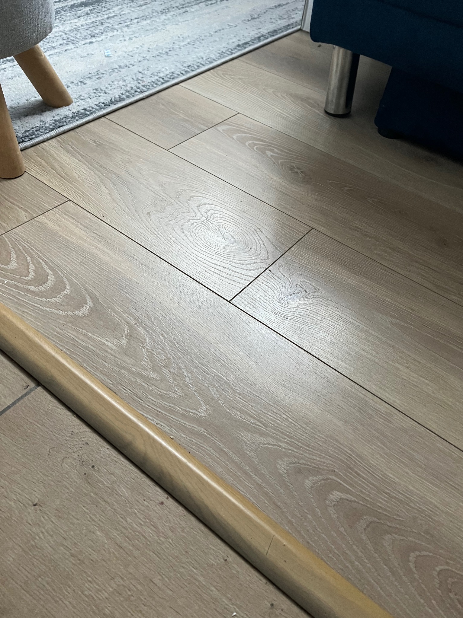 Jasne panele podłogowe z widocznym wzorem drewna, przy listwie przypodłogowej, fragment mebla i dywanu w tle.