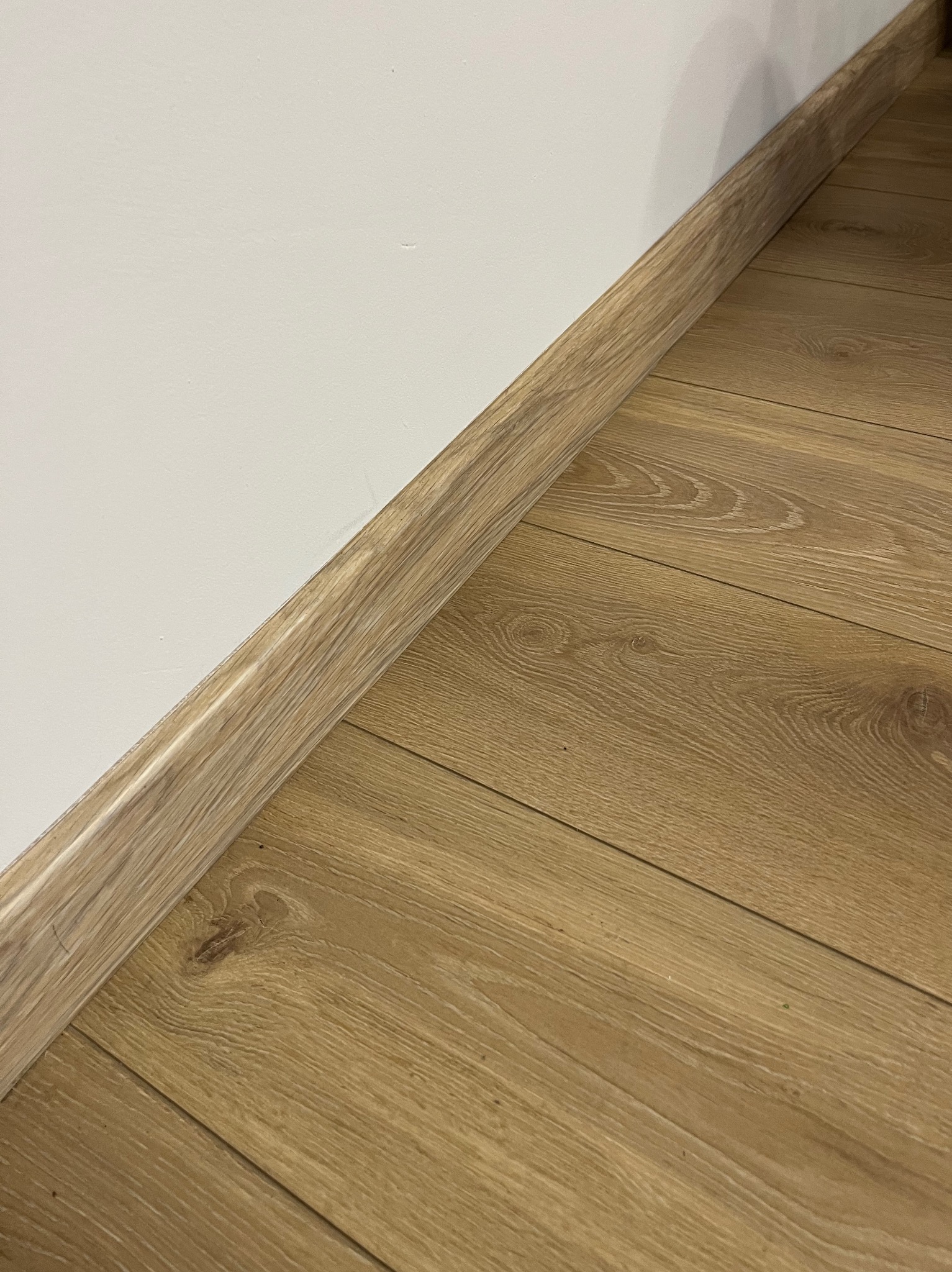 Jasne panele podłogowe z drewnopodobnym wzorem, ułożone przy białej ścianie z dopasowaną listwą przypodłogową.