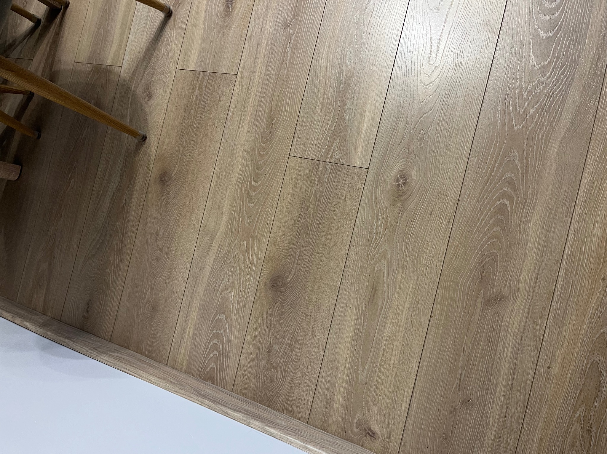 Ułożone panele podłogowe w odcieniach jasnego drewna, widoczne z perspektywy lekko z góry, z fragmentem nogi krzesła w lewym górnym rogu i białą listwą przypodłogową w dolnej części kadru.