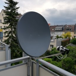 Im-Sat Dortmund - Szara antena satelitarna zamontowana na balkonie bloku mieszkalnego, widok na okoliczne budynki i zieleń, pochmurne niebo w tle.